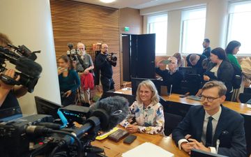 Een vrouw en een man achter een tafel in een zaal met meer tafels en mensen achter hen; op de man en de vrouw zijn camera’s en microfoons gericht.