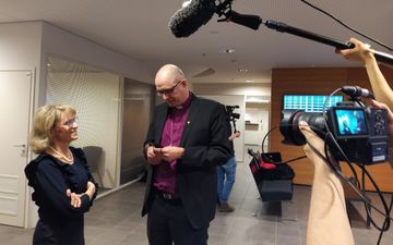 Vrouw in gesprek met veel langere man, terwijl een fotograaf op hen inzoomt