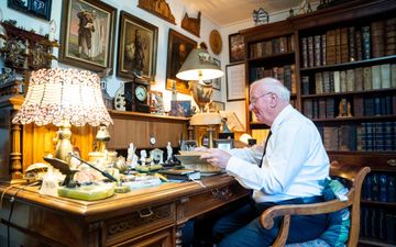 Een oudere man in een wit overhemd achter een bureau, met om zich heen veel kunstvoorwerpen.