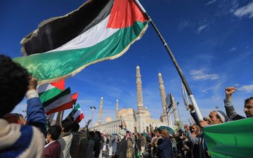 Donkergekleurde mensen met tulbanden en andere hoofddeksels op zwaaien met Palestijnse en Iraanse vlaggen voor een moskee met hoge minaretten en koepels. De lucht is stralend blauw. 