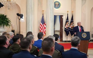 Mensen luisteren in zaal naar man in donker pak die achter spreekgestoelte een toespraak houdt.