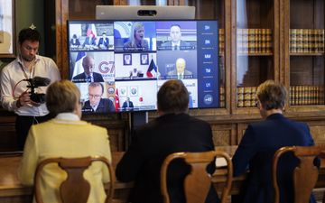Drie mensen zitten achter een tafel voor een groot beeldscherm. Daarachter een boekenkast gevuld met oude boeken. 