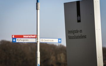 Wit bord met daarop in blauwe letters de tekst ”Immigratie- en Naturalisatiedienst” met op de achtergrond wegwijzers voor vluchtelingen.