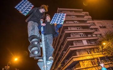 Twee jongelui klimmen ‘s nachts in de paal van een stopilcht en houden borden met”Tisza” erop vast. Op de achtergrond een flatgebouw.