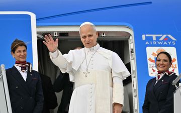Paus Leo, een man in het wit gekleed, zwaait bij een blauw vliegtuig.