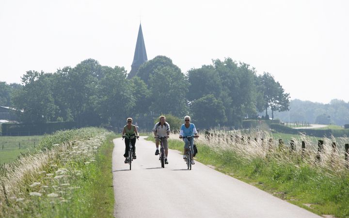 Drie fietsers rijden over een asfaltweggetje over een lage dijk. De foto is van voren genomen. Op de achtergrond zijn bomen te zien met een kerktoren die erbovenuit steekt. De zon staat hoog aan de hemel.