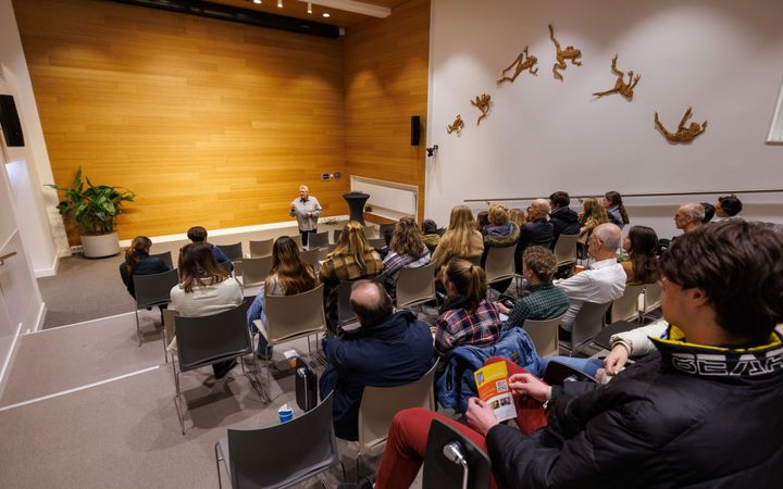 Studenten van allerlei leeftijden in een collegezaal. Op de muur een driedimensionaal kunstwerk van springende kikkers. 