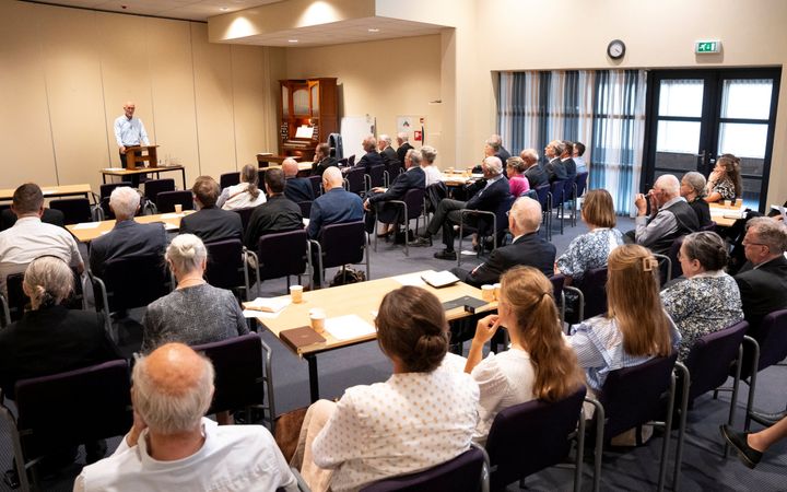 Een zaal vol mensen; ze luisteren naar een man achter een katheder.