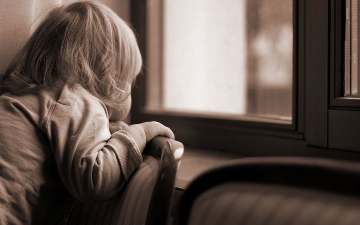 Sepiafoto van een blond kind dat over de leuning van een stoel door een raam staart.