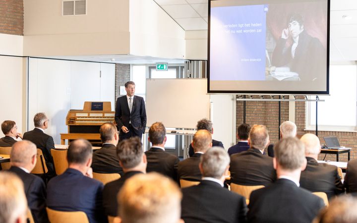 Man in donker pak met stropdas staat voor zaal gevuld met mannen in pak. Op de achtergrond een orgel en beamer.