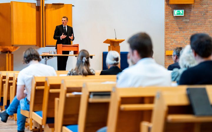Dominee Hoogerbrug, een man in zwart kostuum, staat achter een tafel voorin een kerk, terwijl mensen op kerkbanken naar hem luisteren. 