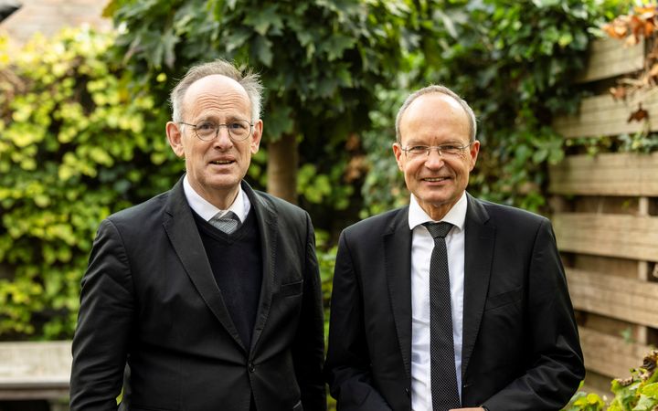 Dominee De Vries en dominee Brons, twee mannen met bril en zwart pak, in een tuin.