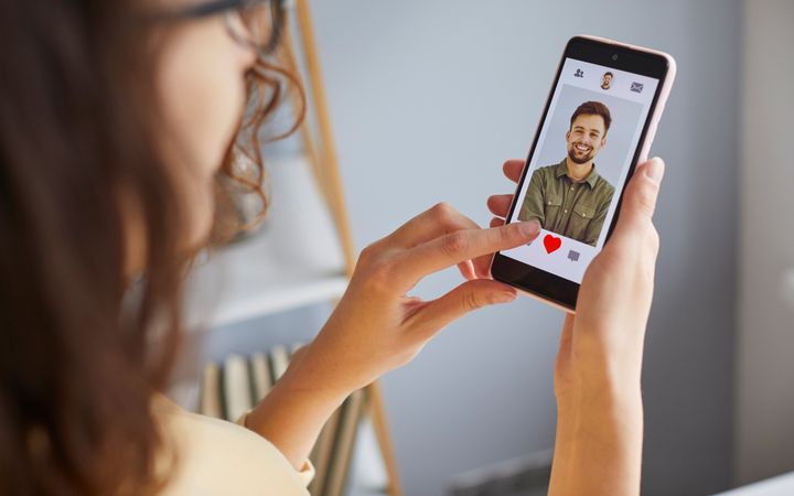 Meisje met bruin haar kijkt op haar telefoon naar een datingapp. Op haar mobieltje is een lachende jongeman zichtbaar.