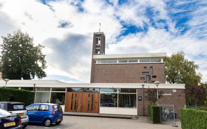 Tamelijk modern kerkgebouw, met een torentje. Op de voorgrond enkele auto’s in parkeervakken.