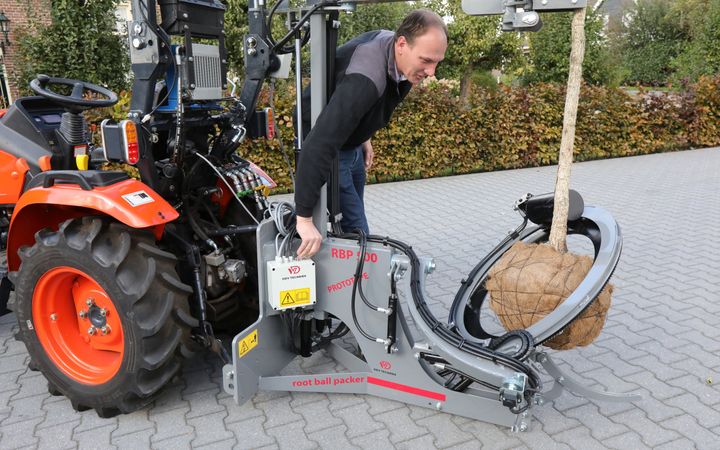 Een man bedient een machine achter een kleine trekker. De machine wikkelt een net rond de kluit van een boom. Om het net zit ijzerdraad.