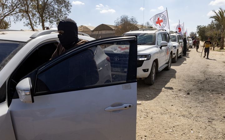 Een colonne witte auto’s met vlaggen van het Rode Kruis in een zanderige omgeving. Vooraan stapt een man met een bivakmuts in een zilverkleurige auto.