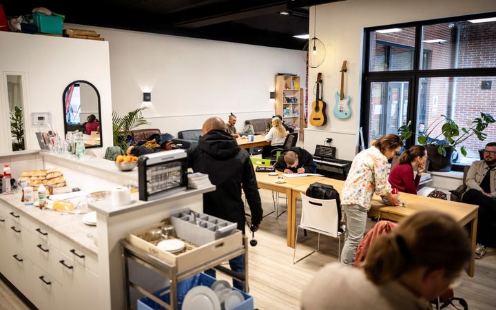 Een grote lichte ruimte met een tiental mensen dat zit of staat. Links een keukenblok, langs de muur drie banken, verschillende gitaren en een keyboard. In het midden een lange houten tafel.