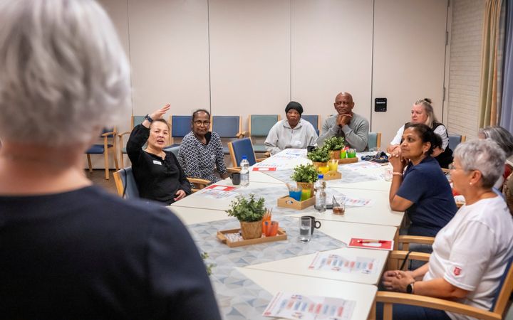Groep mensen zit achter een tafel. Op de voorgrond is een vrouw te zien op de rug. Een van de mensen aan de tafel heeft haar hand opgestoken, de anderen kijken naar haar. 