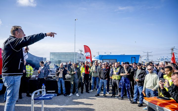 Een groep mannen in werkkleding luistert naar iemand met een microfoon in de hand, die hen toespreekt. Op de achtergrond staat een vlag van vakbond FNV.