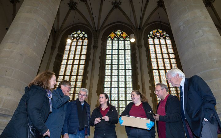 Zeven mensen in gesprek, met achter zich de metershoge glas-in-loodramen.