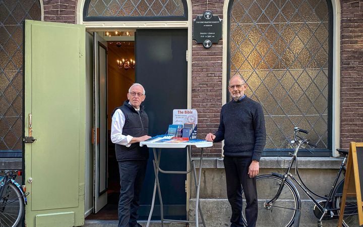 Twee mannen staan voor de gevel van een kerkgebouw. Tussen hen in een statafel met evangelisatiemateriaal.