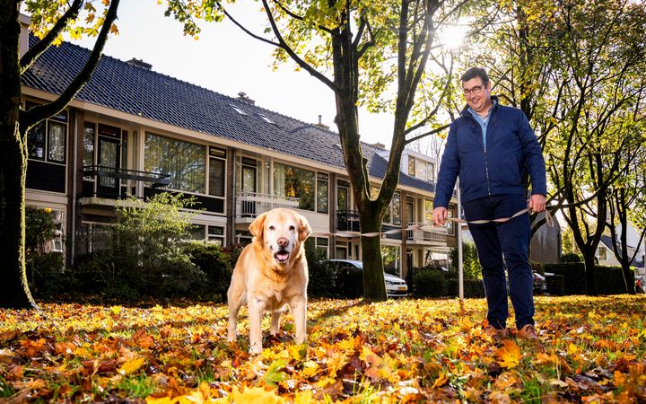 Een man met bril en een blauwe jas aan staat met een hond aan de riem staat buiten op een grasstrook met veel herfstbladeren. Op de achtergrond staan rijtjeshuizen.