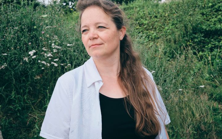 Vrouw met wit-zwarte kleding en lange donkerblonde haren kijkt weg naar links. Op de achtergrond een groen landschap.
