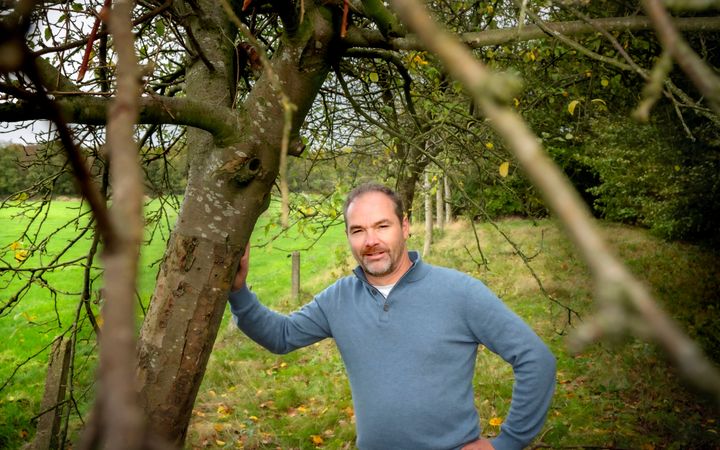 Een man met een lichtblauwe trui aan leunt met zijn arm tegen een boom. Op de achtergrond grasland met een hekwerk.