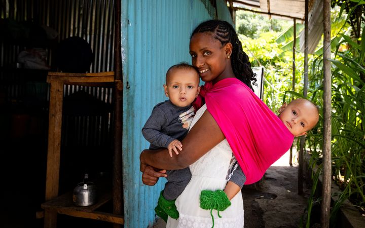 Jonge moeder met jurk draagt een tweeling. De ene baby zit in een draagdoek op haar rug. De andere draagt ze op haar buik. 