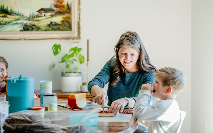 Vrouw van middelbare leeftijd in grijsblauwe jurk snijdt brood voor een jongetje dat aan tafel zit. Op de tafel staan bekers en etenswaren.