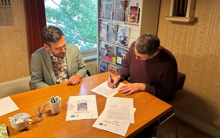 Twee mannen zitten aan een tafel en zijn bezig met het ondertekenen van een overeenkomst.