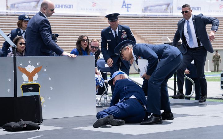 Man in formele kleding met petje op krabbelt overeind en wordt daarbij geholpen door toegesnelde mannen.