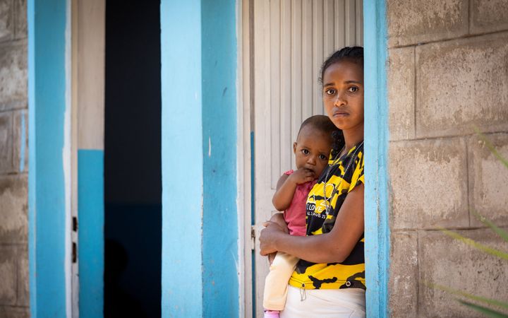 Meisje staat in deuropening met en kijkt verdrietig in de lens. Ze heeft haar dochter op haar heup. 