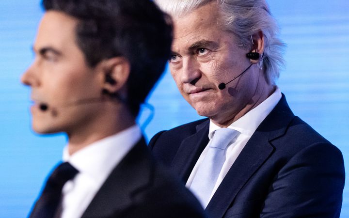 Een gespannen kijkende man met geblondeerd haar kijkt langs een man met donker haar wat grijs begint te worden. Allebei dragen ze een pak.