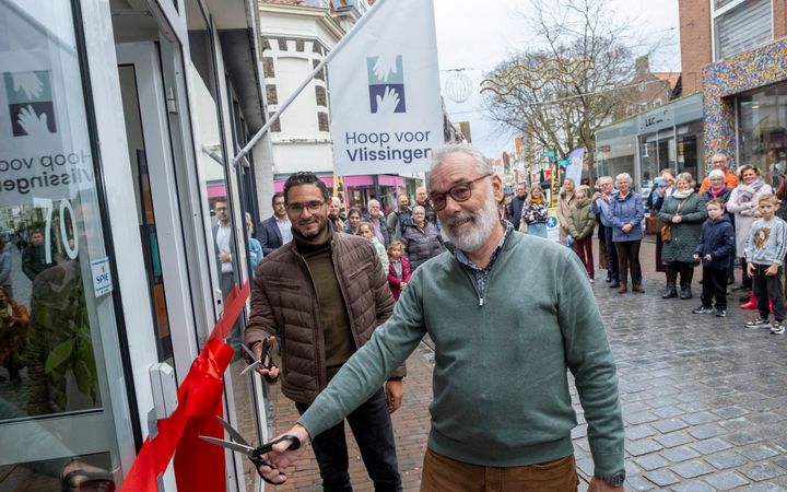 Twee mannen houden een schaar bij een rood lint voor de ingang van een pand. Op de achtergrond kijken mensen toe.