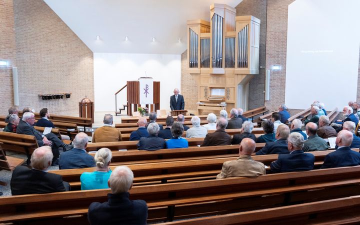 Een kerkzaal met vooral mannen in banken; ze luisteren naar dominee De Graaf voorin.