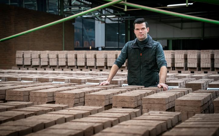 Man met donker haar en donkere bodywarmer staat tussen stapels baksteunen waar hij met beide handen op leunt. 
