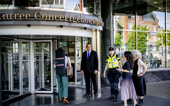 Twee dames in lange rok lopen naar een draaideur waarboven staat: Entree Concertgebouwplein. Ook een vrouw met een koffer loopt naar binnen. Bij de deur staat een man in een donker pak en een politieagente.