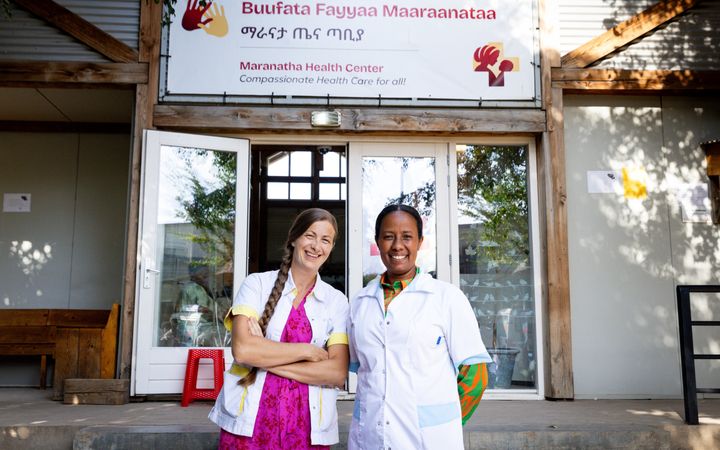 Twee vrouwen staan in witte jassen voor de deur van de Maranathakliniek, de vrouw links heeft een lange vlecht. 