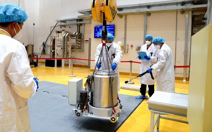 Mensen met witte labjassen aan, met mondkapjes voor en blauwe haarnetten op hun hoofd verplaatsen een metalen cilinder met een ingebouwde kraan. 