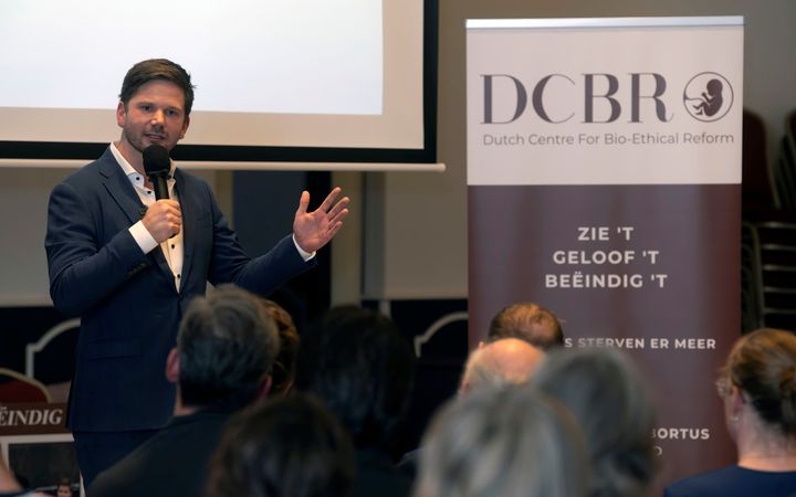 Man in blauw pak en witte blouse spreekt in microfoon naast banner met letter DCBR erop.