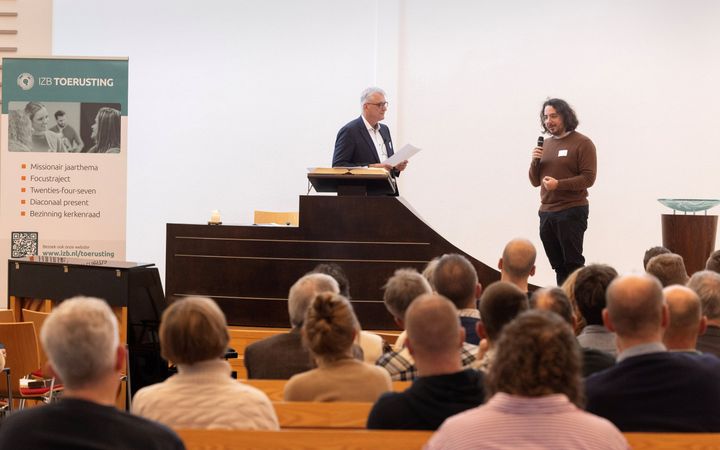 Man met een trui, vrij lang haar, doet zijn verhaal via de microfoon. Links ds. Van der Graaf.