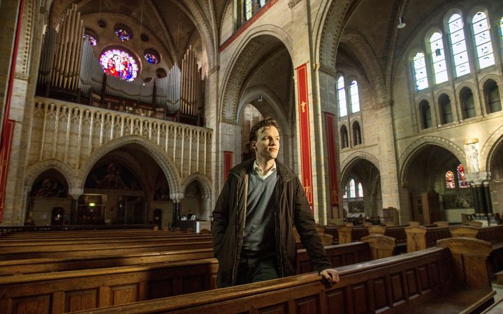 Een jongeman staat in een grote kerk tussen de kerkbanken en kijkt de kerkruimte in. Op de achtergrond is het brede front van een groot pijporgel te zien.