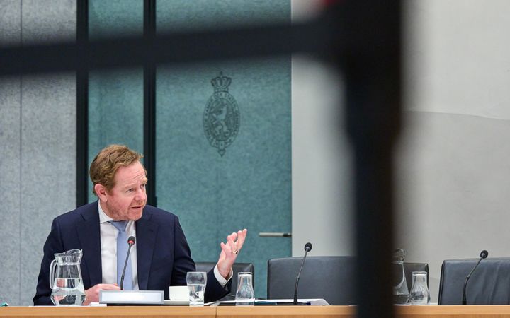 Man in pak zit in een commissiezaal van de Tweede Kamer en gebaart met zijn hand.