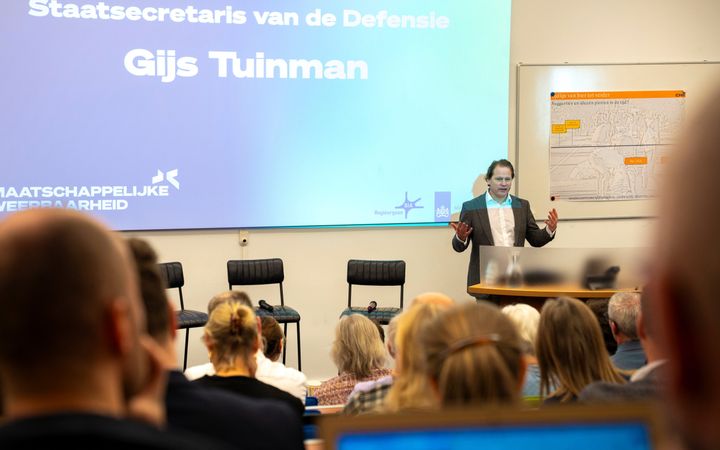 Een man staat achter een desk voor een groep mensen in een collegezaal.