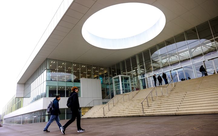 Twee jongeren met rugzakken lopen richting een brede trap. Bovenaan staan anderen jongeren op hun mobiele telefoon te kijken. 
