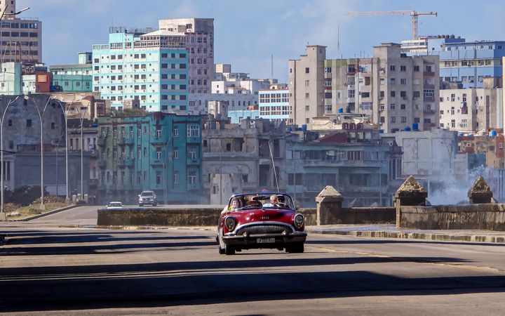 Een rode oldtimer rijdt over een vrijwel lege weg. In de verte zijn gebouwen in slechte staat zichtbaar. 