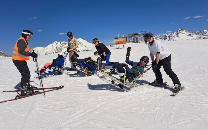 Foto op een piste. Drie mensen staan achter een zitski, een soort buggy op latten. Links staat een meneer op normale ski’s. De lucht is blauw, op de achtergrond zijn de bergen te zien en een stoeltjeslift.