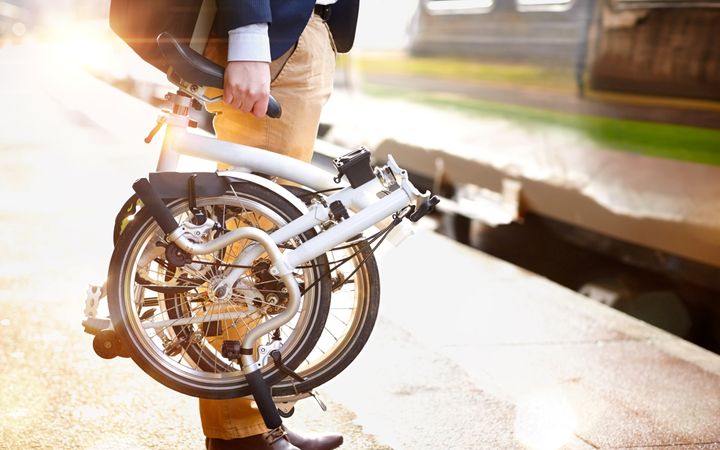 Een man met een opgevouwde vouwfiets in zijn rechterhand wacht op een perron op de trein.