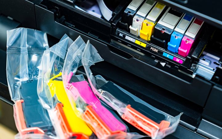 Inktcartridges in plastic verpakkingen in de kleuren roze, blauw, zwart en geel liggen voor een printer waarvan de kap openstaat. 